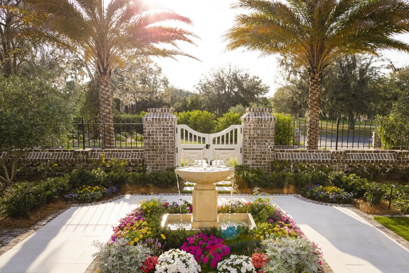 Garden fountain surrounded by flowering beds, palms and a white gate