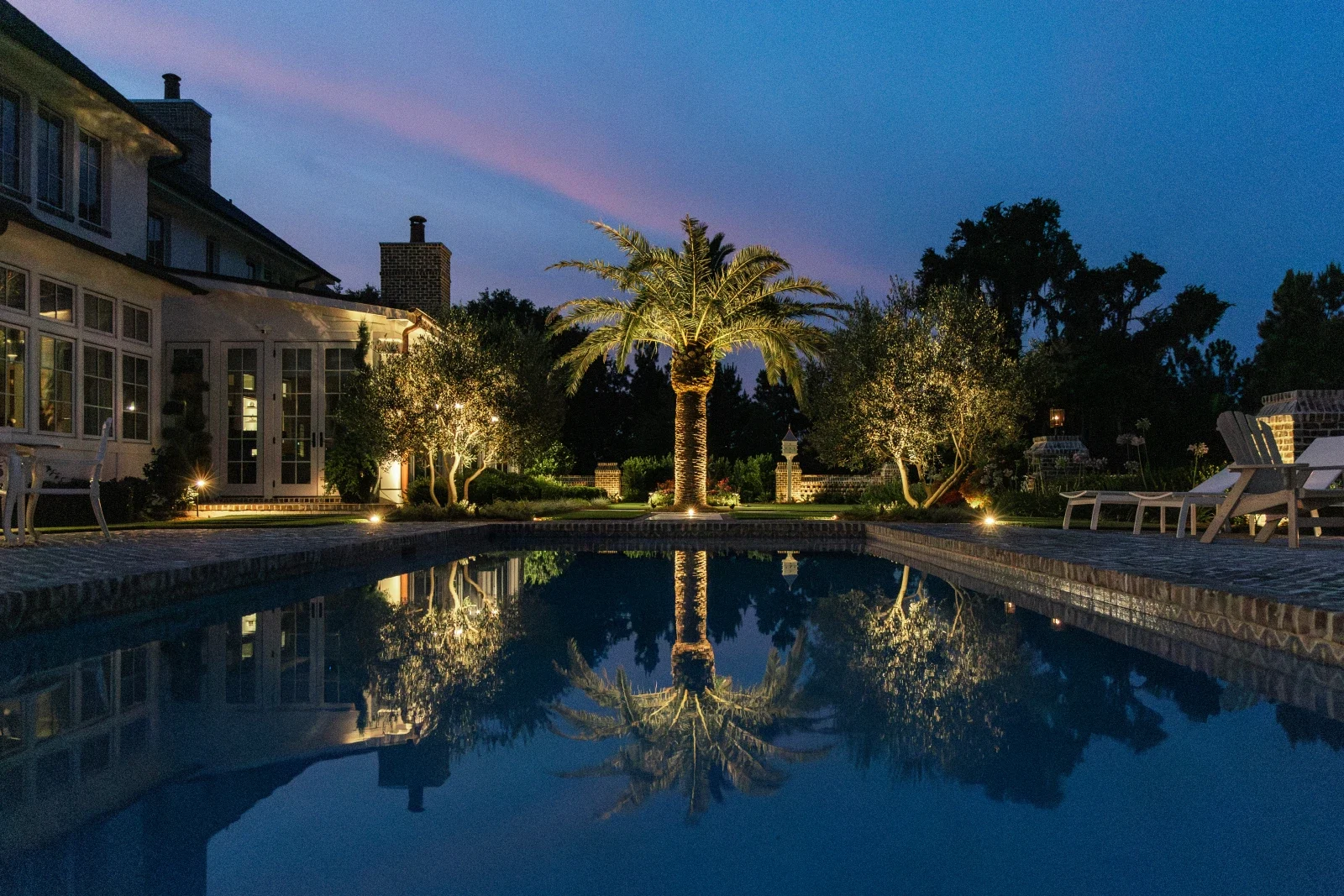Warm landscape lighting illuminating palms, planting and a pool at dusk